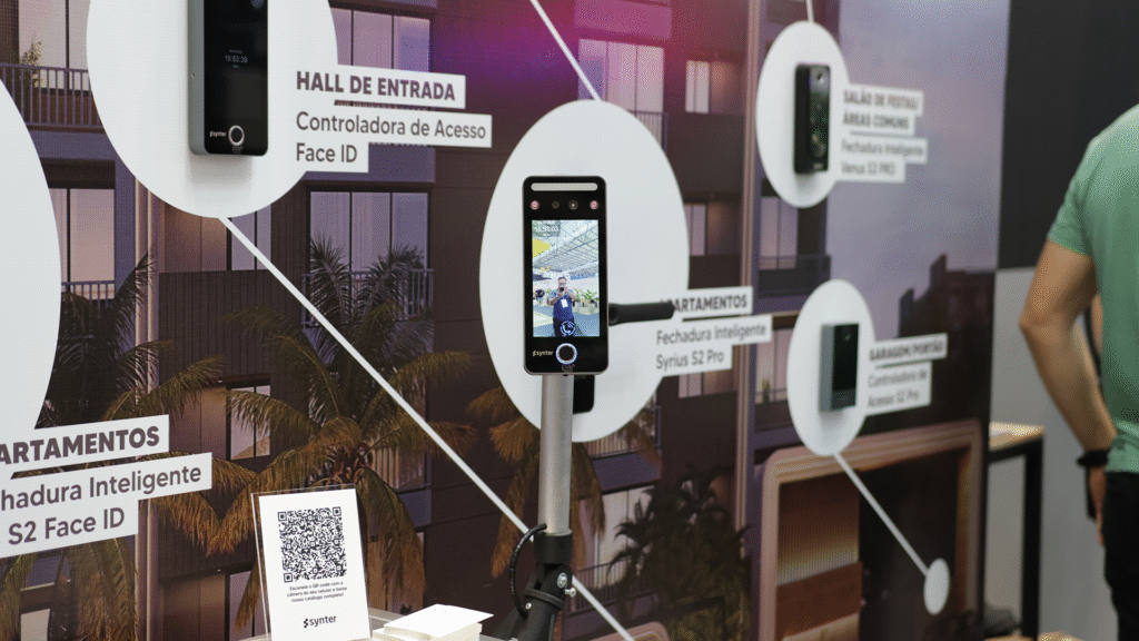 A imagem mostra um detalhe do painel do Synter Access, parte do estande da Synter, apresentando uma simulação de um edifício com a aplicação prática das soluções inteligentes da marca para controle de acesso em diferentes áreas do condomínio.
Os produtos são destacados com imagens reais posicionadas em frente ao painel, e conectados visualmente por linhas que apontam para os respectivos locais de instalação no prédio ilustrado. Os dispositivos e suas localizações são:
Hall de Entrada: Controladora de Acesso Face ID, um equipamento com reconhecimento facial, mostrado em funcionamento ao centro da imagem, com tela ativa exibindo a imagem da feira ao fundo.
Apartamentos: Fechadura Inteligente Syrius S2 Pro
Salão de Festas / Áreas Comuns: Fechadura Inteligente Venus S2 Pro
Garagem / Portão: Controladora de Acesso S2 Pro
O conjunto visual é organizado, didático e tecnológico, permitindo que os visitantes compreendam rapidamente como os dispositivos Synter Access se integram para oferecer segurança, automação e praticidade em empreendimentos residenciais.