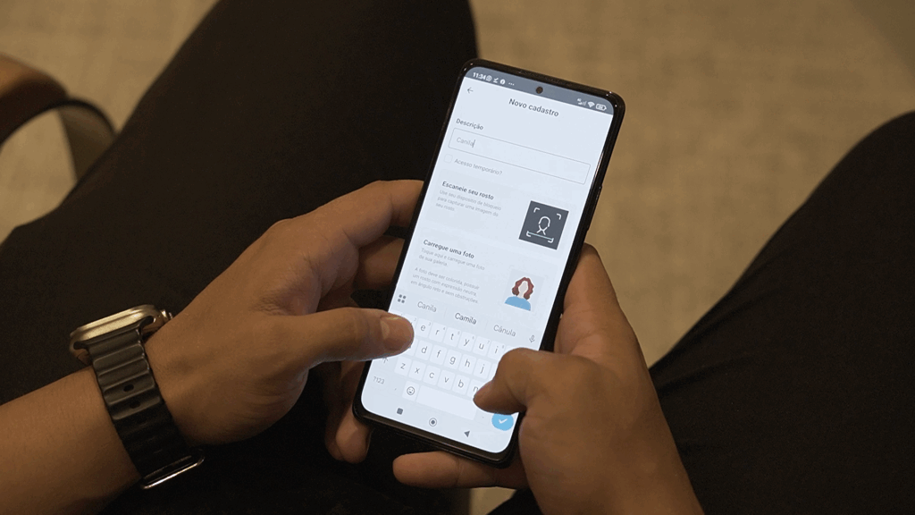 Pessoa segurando um smartphone com as duas mãos, digitando em um campo de cadastro no aplicativo. A tela mostra a seção 'Novo cadastro', com campos para descrição, foto e reconhecimento facial.